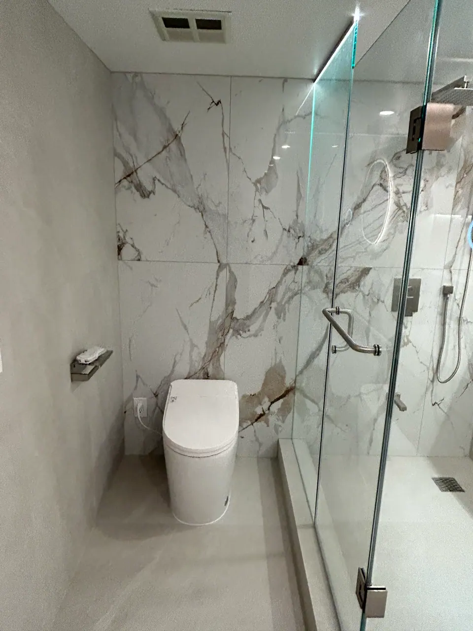 The book-matched Calacatta wall in the finished ensuite running full height behind a concealed-tank toilet, with the glass enclosure and its chrome pull at right and soft grey large-format panels opposite.