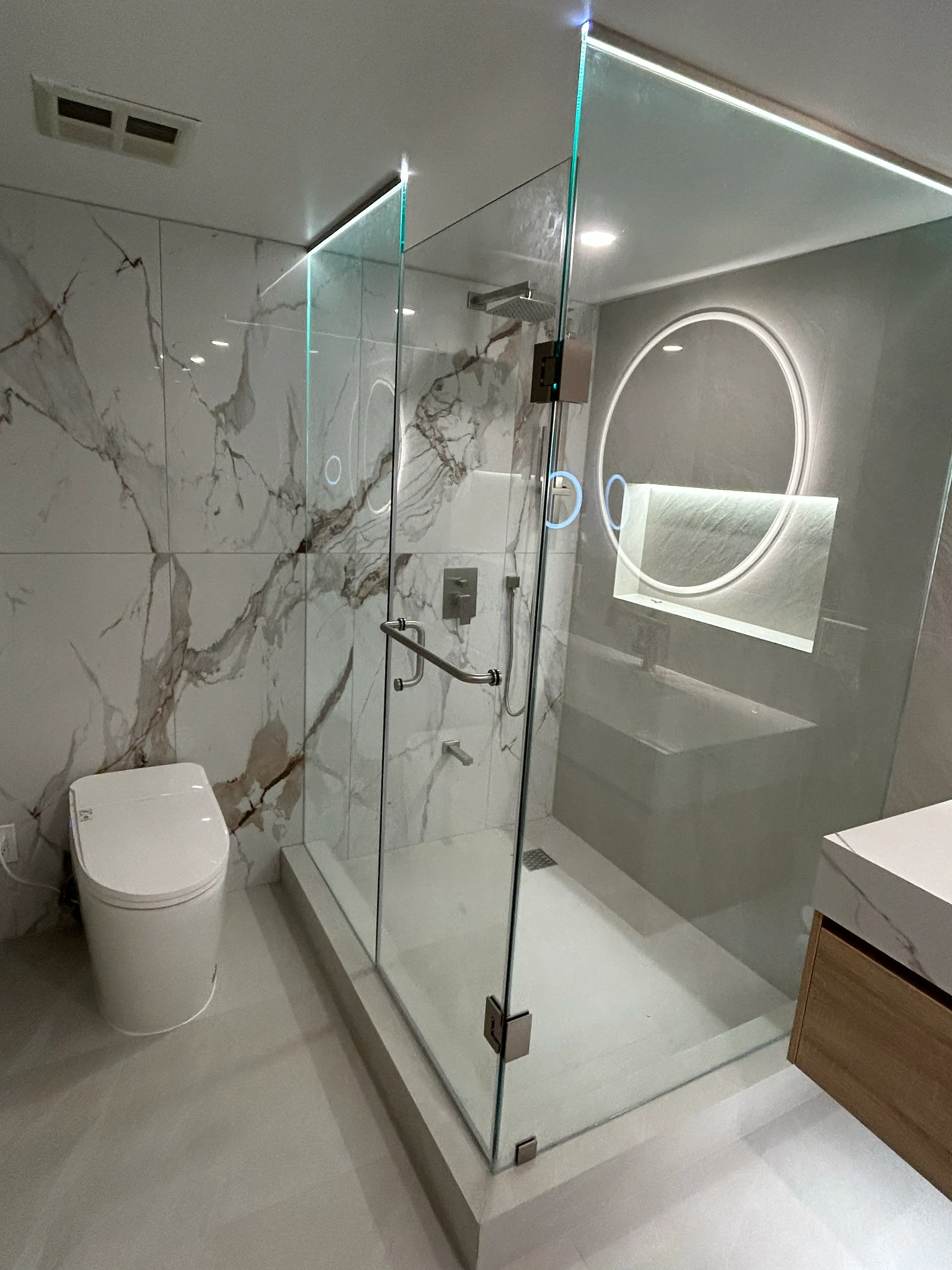 The finished ensuite: a frameless glass shower enclosure against a book-matched Calacatta wall, a backlit round mirror over a floating oak vanity with an integrated stone basin, and a concealed-tank toilet on the left.