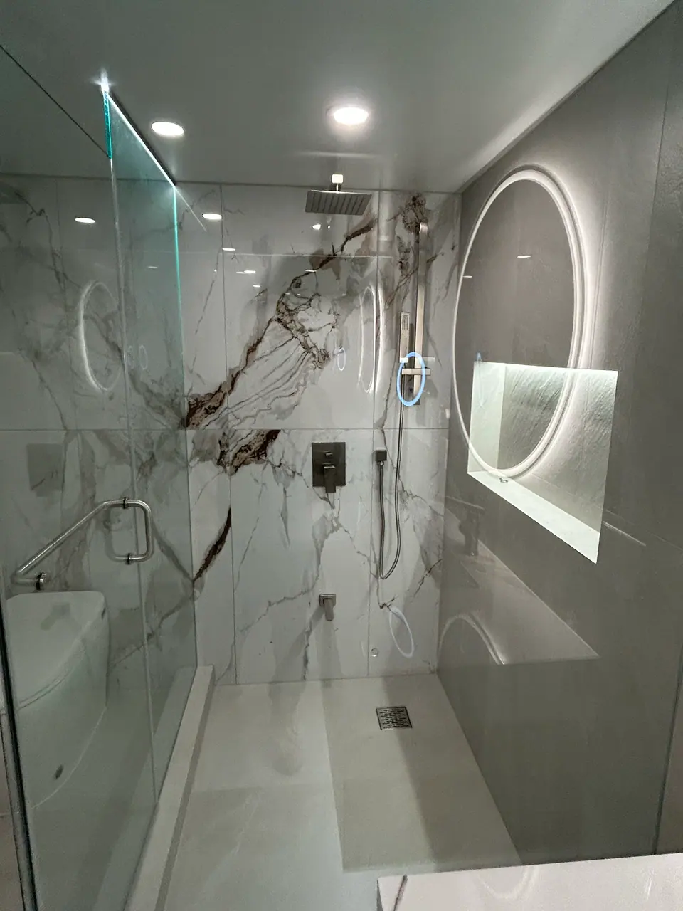 Inside the finished shower: a square rain head, a hand shower on a slide bar, and a flush mixer plate set against the book-matched Calacatta, with the backlit mirror and niche visible through the glass beyond.