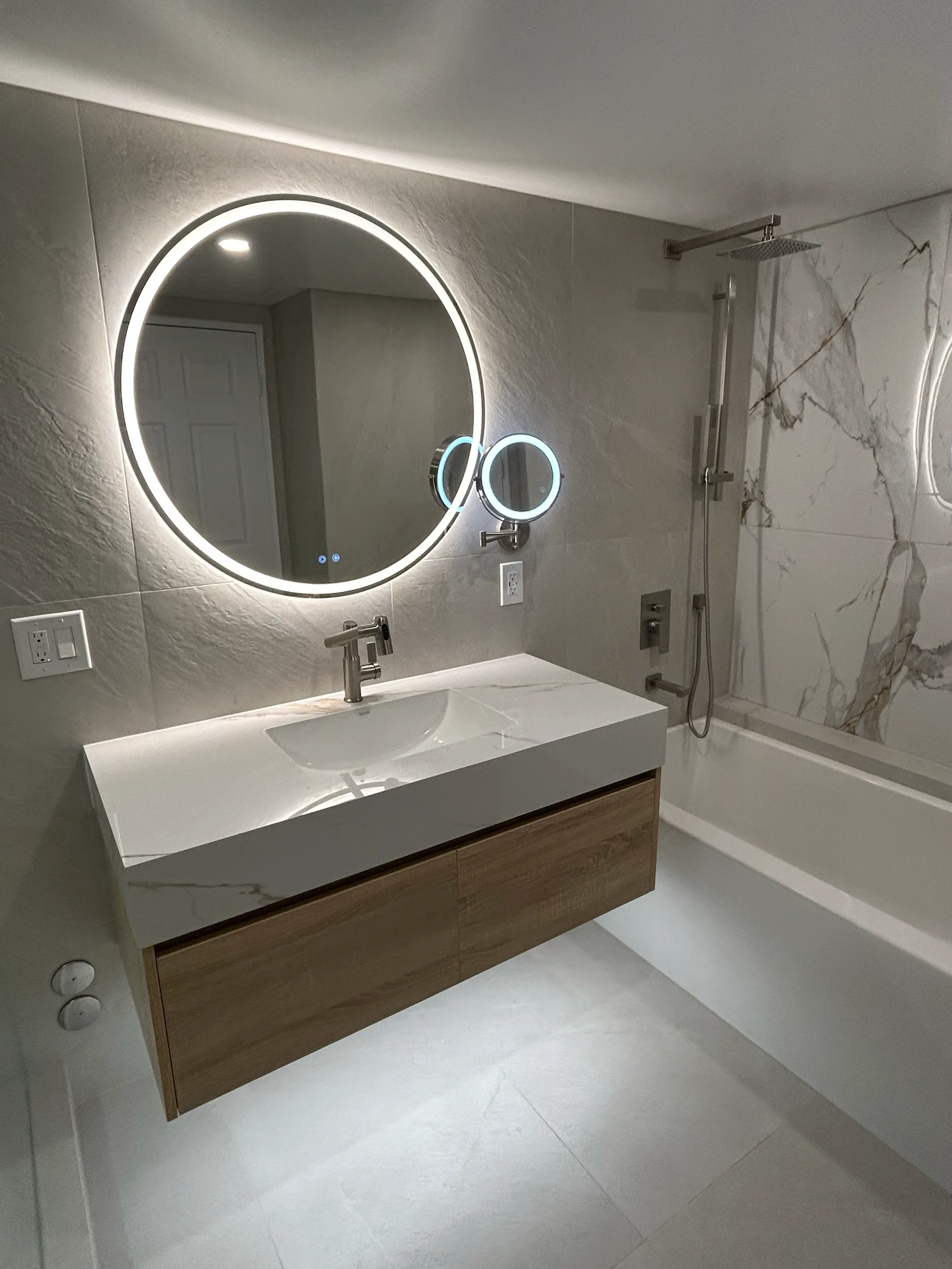 The finished second bath: a floating oak vanity with an integrated stone basin under a backlit round mirror, a chrome magnifying mirror alongside, and the tub with its rain head beyond.