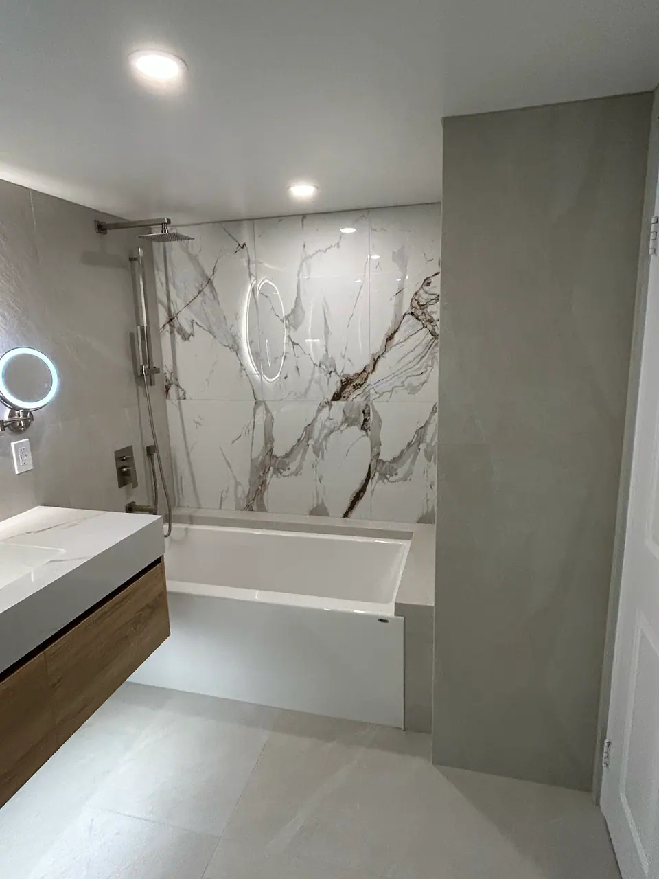 The finished second bath wide: the tub set beneath a book-matched Calacatta panel with a wall-mounted rain head and slide bar, the oak vanity at left, and a full-height greige partition to the right.