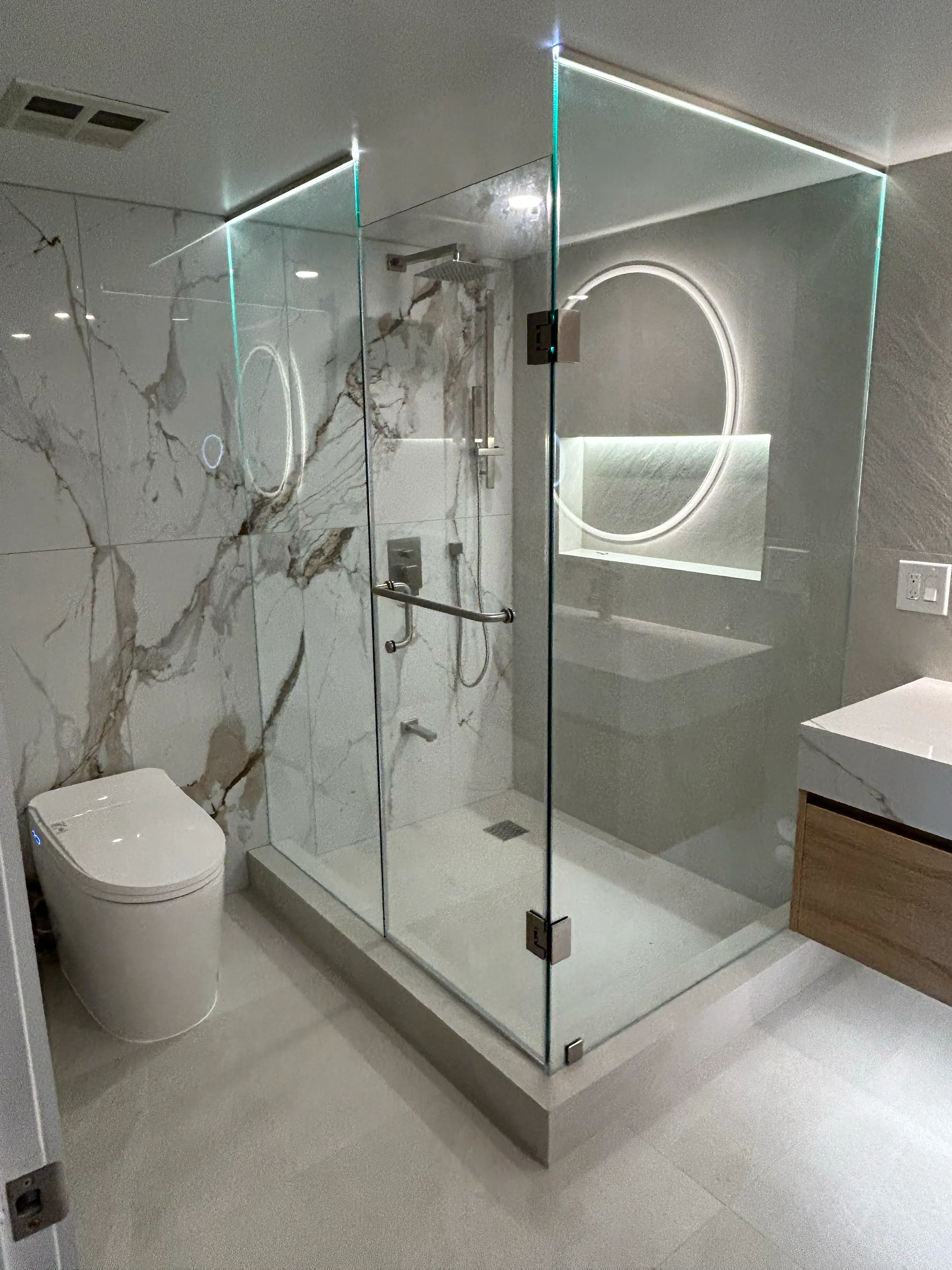 The finished ensuite from the doorway: the glass enclosure reads as a clear box set into the room, with the book-matched wall behind it, the vanity at right and the backlit mirror reflected in the glass.