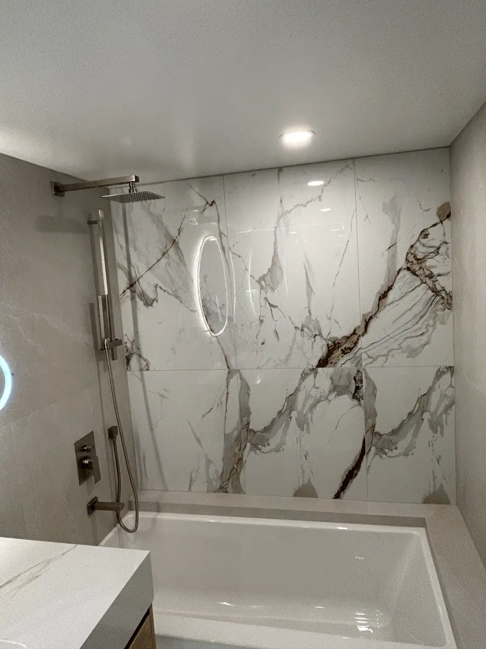 The tub wall finished: a book-matched Calacatta panel running full height above the tub, with a square rain head on a wall arm, a hand shower on a slide bar, and a flush mixer plate.