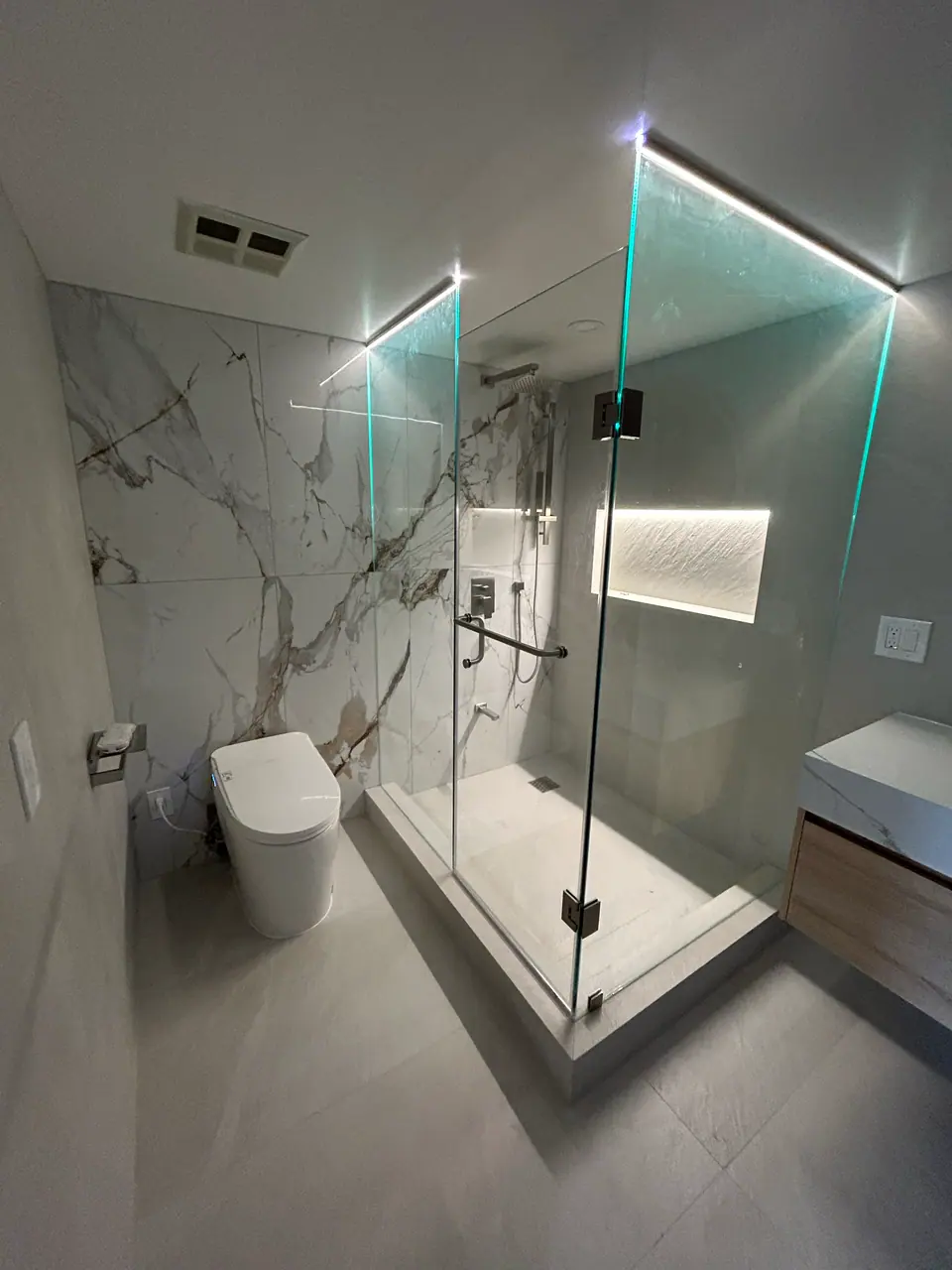 The finished ensuite lit at night: the exposed edges of the frameless glass catch the light and glow along their length, and the recessed niche in the shower is lit from within.