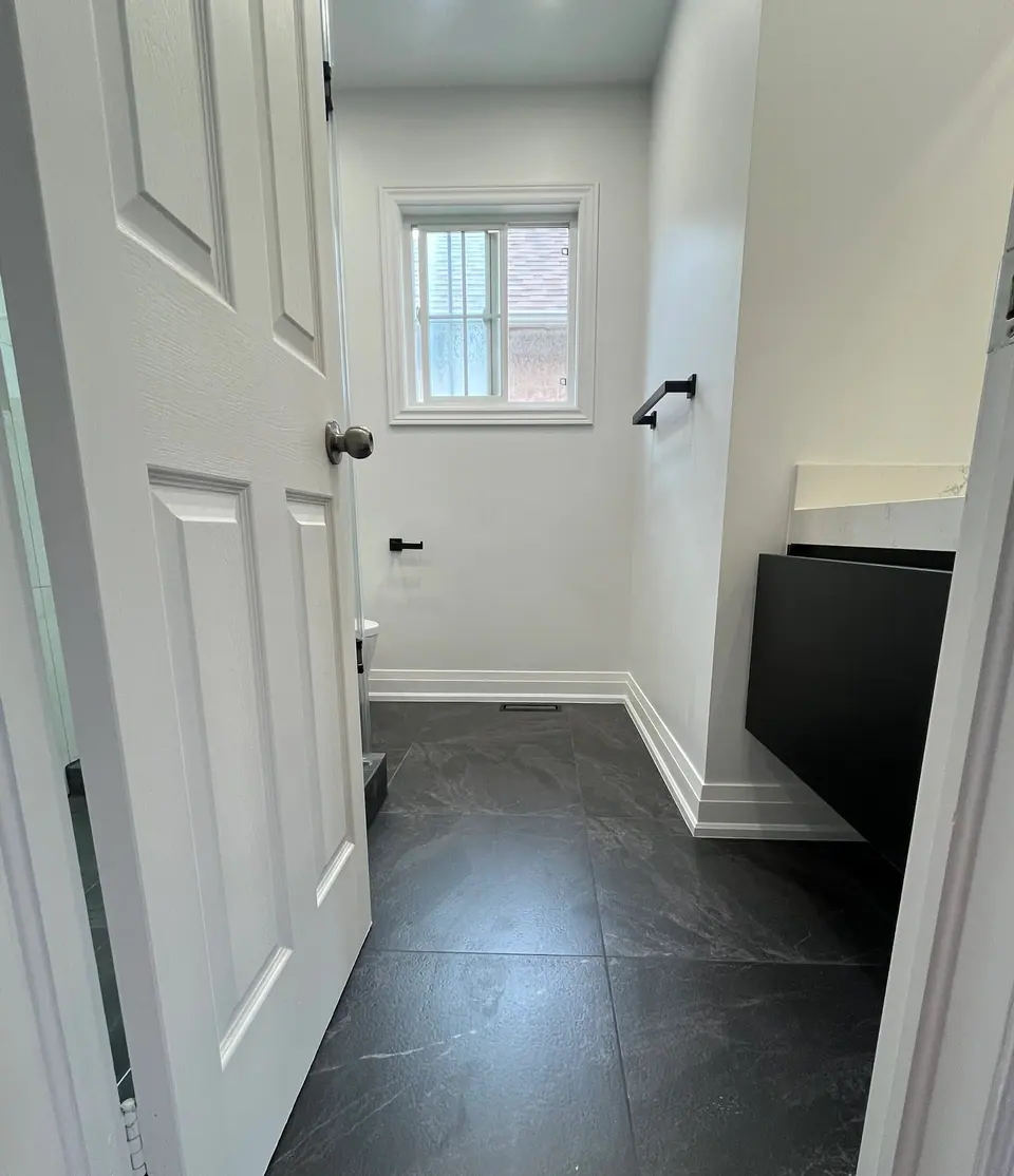 The finished bathroom from the doorway: large slate-look porcelain laid across the floor, white walls and crisp painted baseboard, the original window reinstated, a matt black towel bar, and the edge of the black floating vanity with its white stone top.