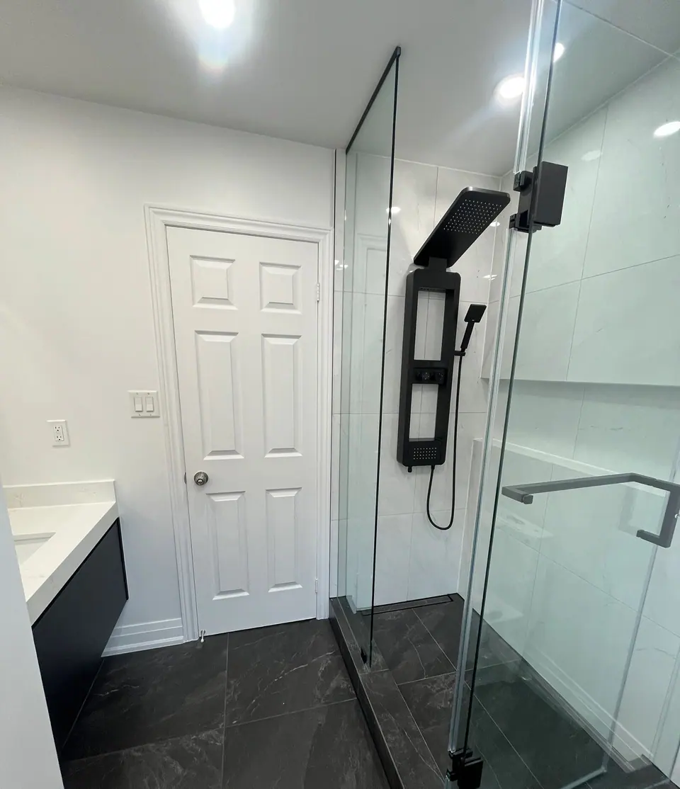 The finished walk-in shower: frameless glass on a slate-look base, a matt black shower panel with a rectangular rain head and hand shower, marble-look porcelain running full height, and a recessed shelf at the far end.