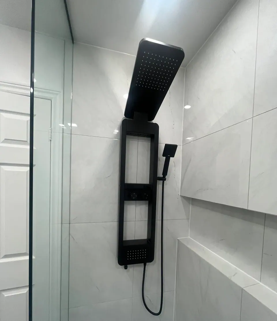The shower panel close up: a matt black column with a wide rain head cantilevered from the top, an integrated hand shower on a coiled hose, and a stepped porcelain shelf running along the return wall.