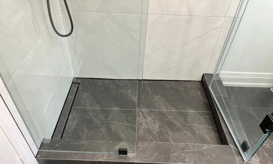 The shower floor finished: slate-look porcelain falling to a linear drain set tight against the glass, a mitred curb capped in the same stone, and the frameless panel landing on it with a flush floor clamp.