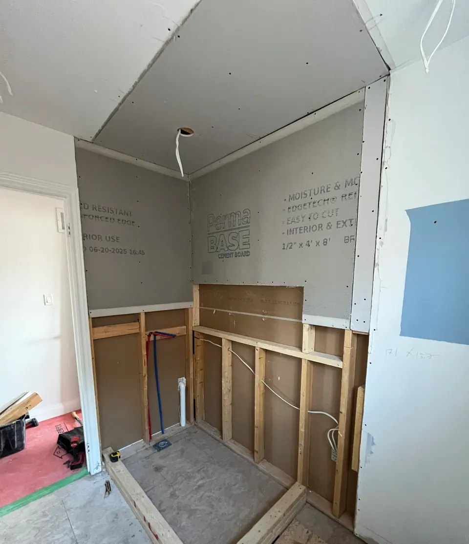 The shower rebuilt in structure: cement board fixed across the back and side walls with the manufacturer print still on it, timber studs and horizontal blocking exposed below, supply lines run in red and blue, and a new curb framed out on the subfloor.