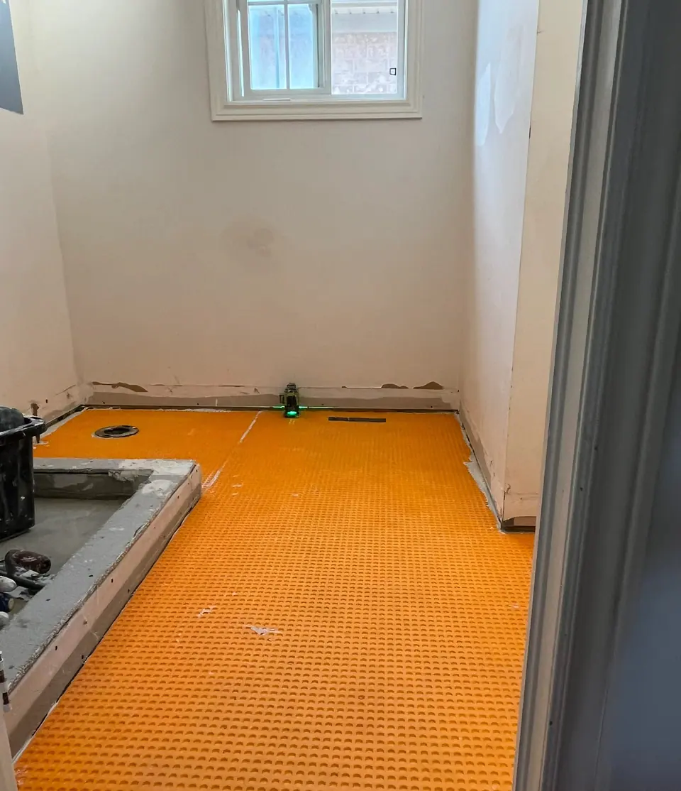 The floor prepared for tile: orange uncoupling membrane bonded across the whole bathroom floor, a green laser line struck along the base of the wall, the shower curb formed in mortar, and the drain body set flush.