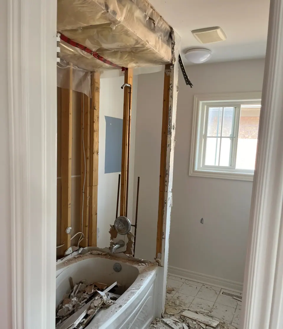 The same bathroom stripped: the tub filled with broken tile and plaster, the wall behind it opened to bare studs and copper supply lines, polythene sheeting taped across the ceiling, and debris across the remaining floor tile.