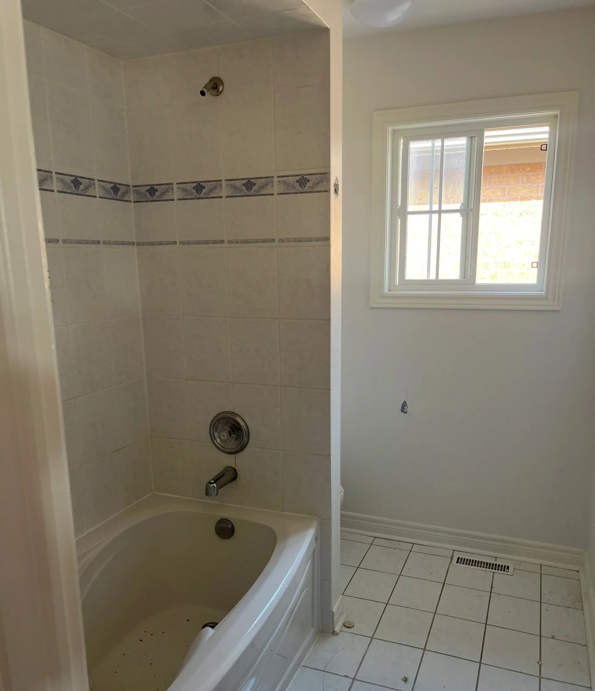 The bathroom before any work: a moulded acrylic tub in a four-inch tile alcove, a blue floral border running at two heights, a chrome pressure-balance valve, and white square floor tile meeting a return-air grille in the corner.