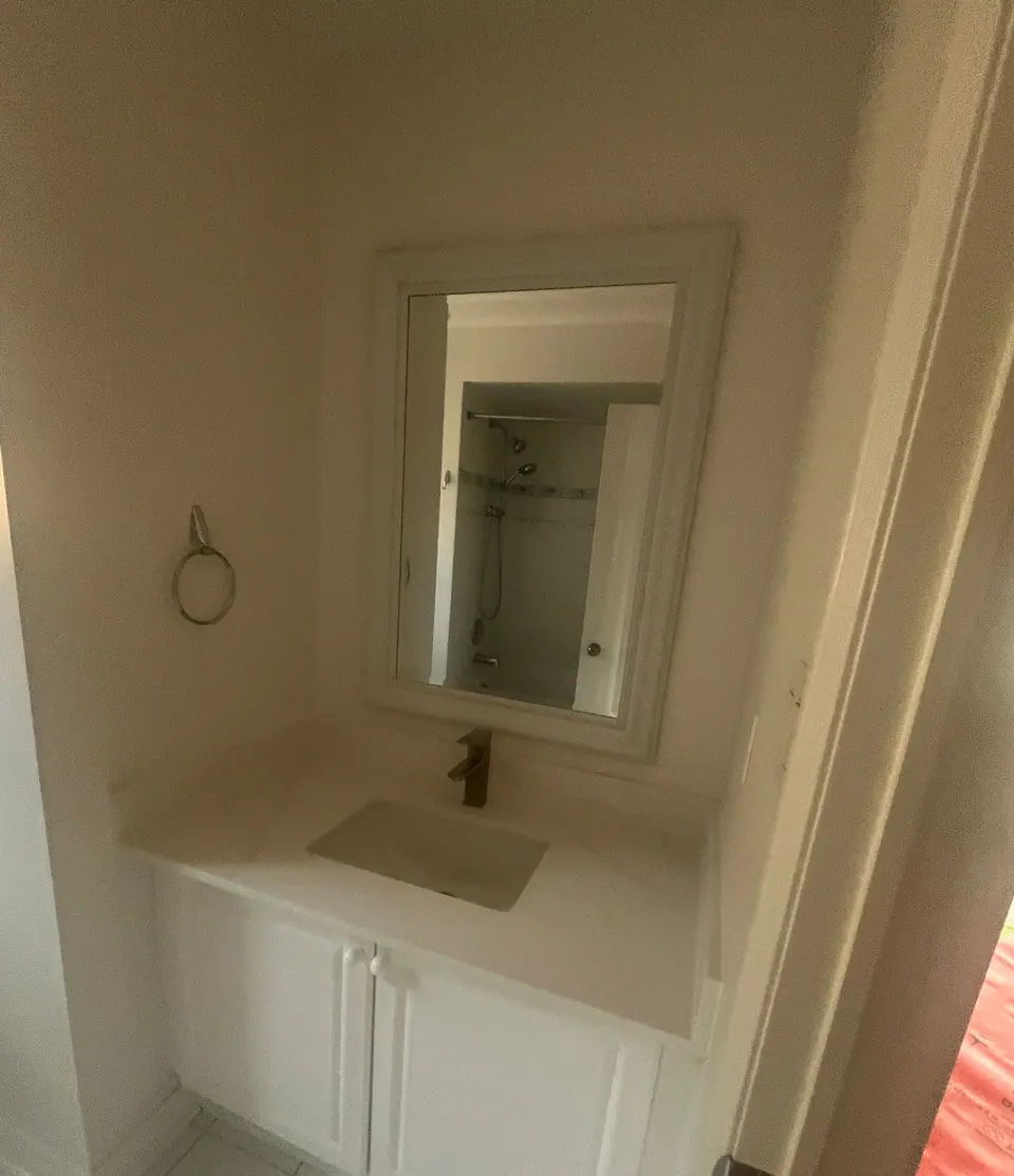 The original vanity nook: a small white raised-panel cabinet under a moulded stone top, a framed mirror set into the recess, a chrome towel ring, and white square floor tile.