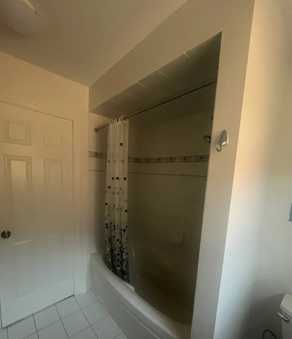 A second original bathroom in the same house: an alcove tub behind a patterned shower curtain, a dark mosaic border at chest height, a chrome shower arm, and white square tile underfoot.