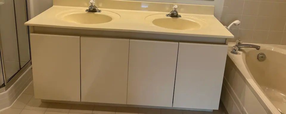 The original vanity before demolition: a one-piece cultured marble top with two moulded basins, cream laminate doors, and a chrome deck-mount faucet at each bowl.