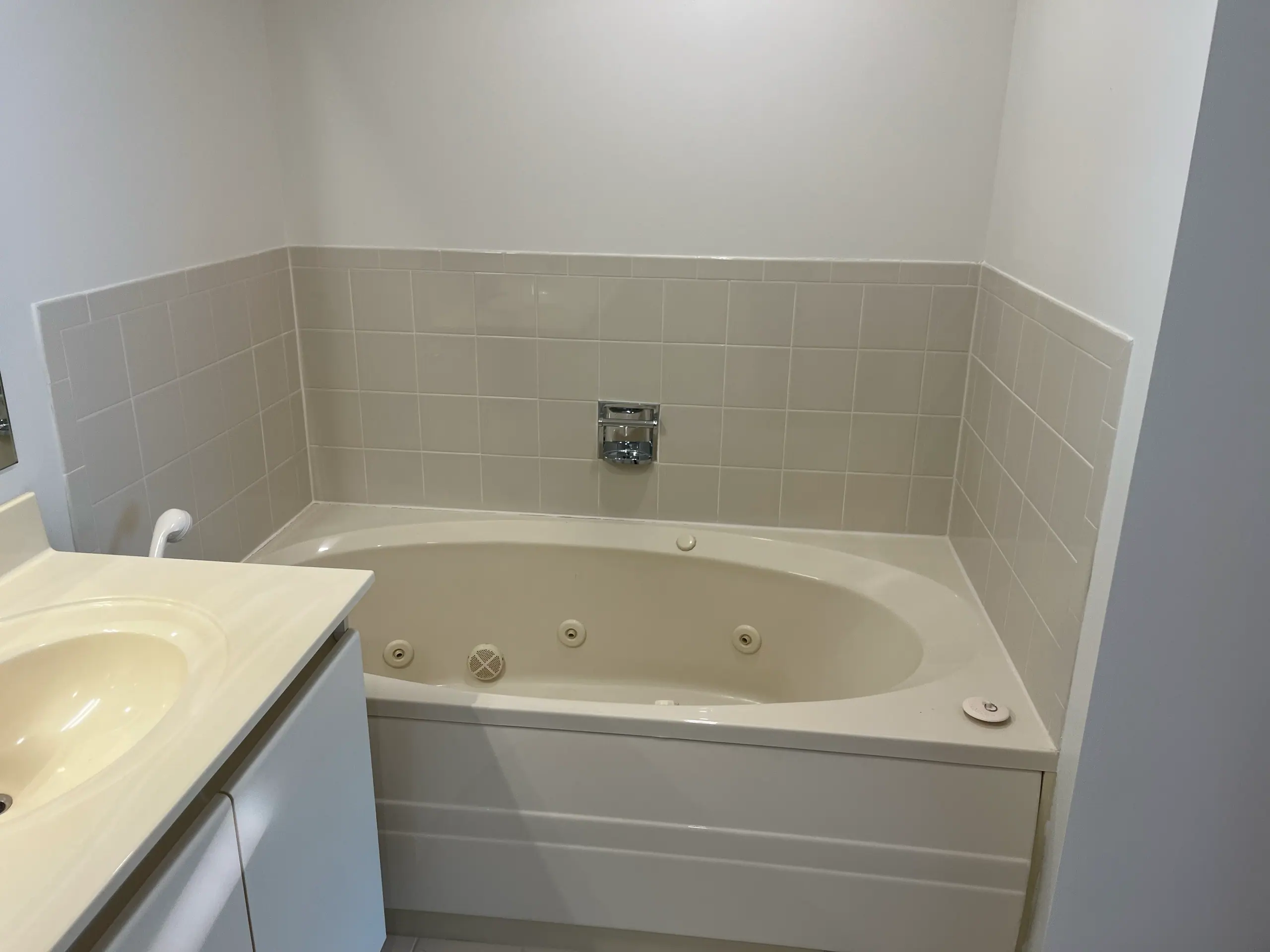 The original corner jetted tub set into a tiled surround, with a small square soap niche and a glass block accent. Four inch tile runs to a bullnose cap partway up the wall.