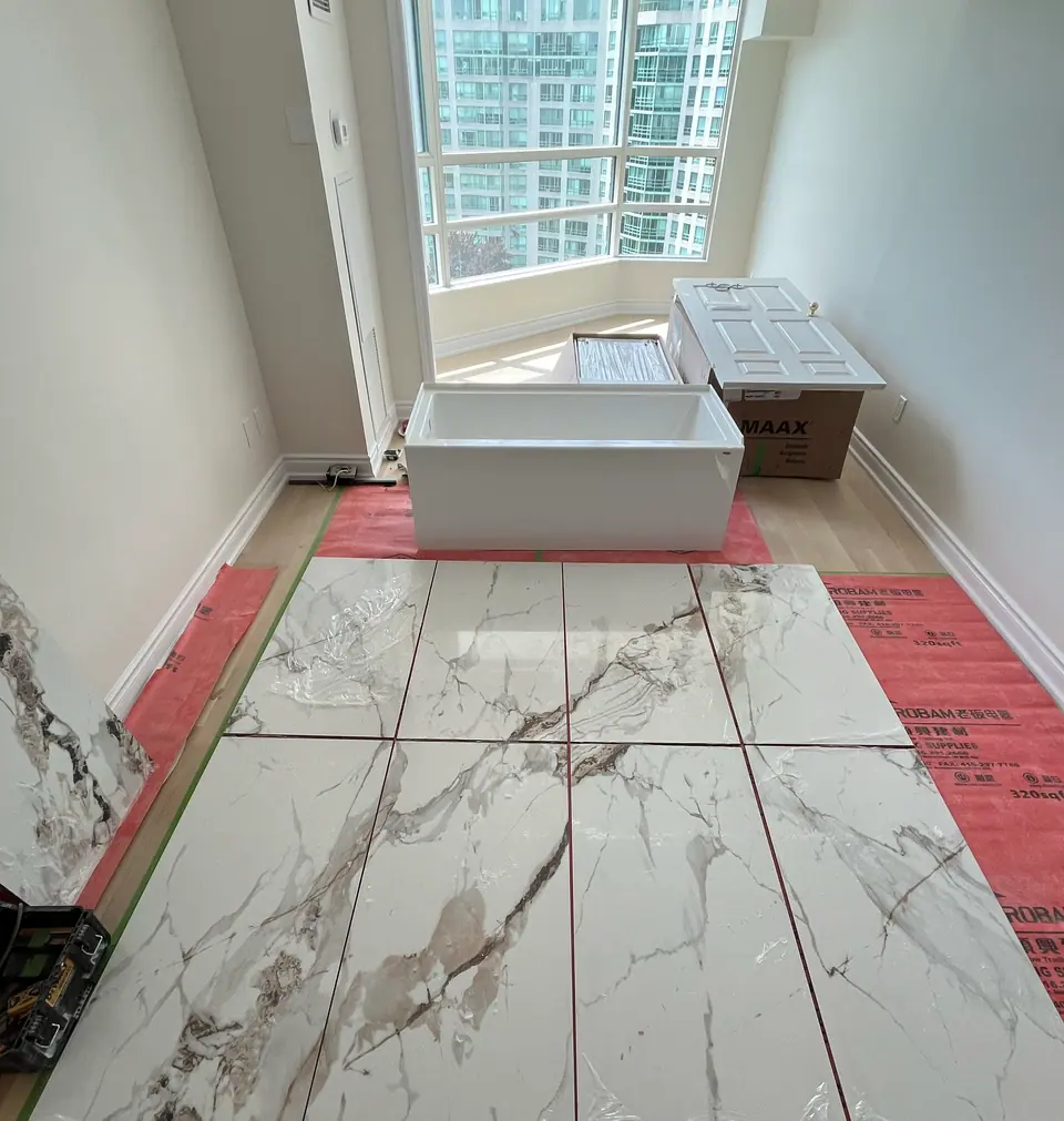 Six large-format porcelain panels dry-laid across the floor in daylight, sequenced so the grey and gold veining runs continuously from one panel to the next. A new alcove tub sits unboxed by the window.