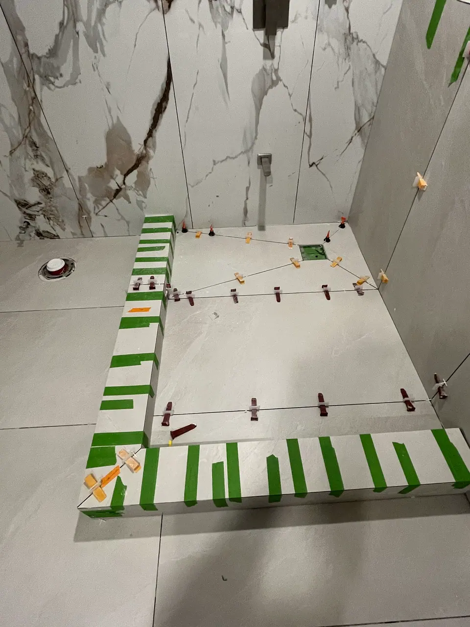 Looking straight down onto the shower curb: the mitred porcelain return is taped along its length, and a string line runs corner to corner across the pan with leveling clips holding the fall true toward the drain.