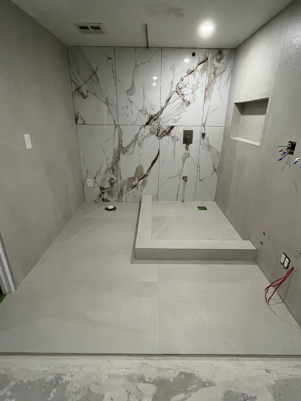 The shower with tiling complete and clips removed: the book-matched feature wall runs floor to ceiling behind a low tiled bench, flanked by wide-format grey panels with joints kept tight and level.
