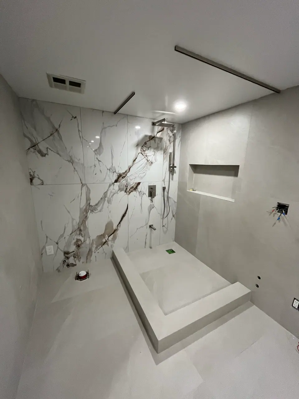 The shower with fixtures set but no glass yet: a ceiling-mounted rain head, a slide bar and handset, and a flush square mixer plate against the book-matched wall. Two concealed lighting channels run the ceiling.