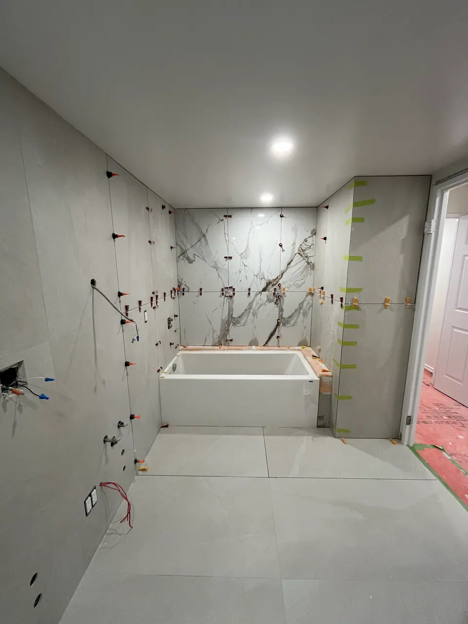 The second bath with the alcove tub in place and the surround being tiled: a book-matched porcelain panel centred above the tub, grey panels running out to either side, clips still holding the joints.